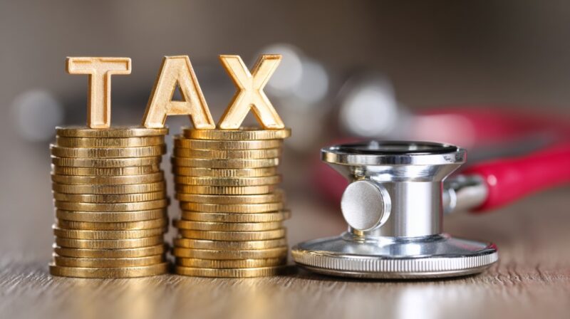 Stacks of gold coins spelling "TAX" beside a stethoscope, representing the link between taxation and healthcare funding